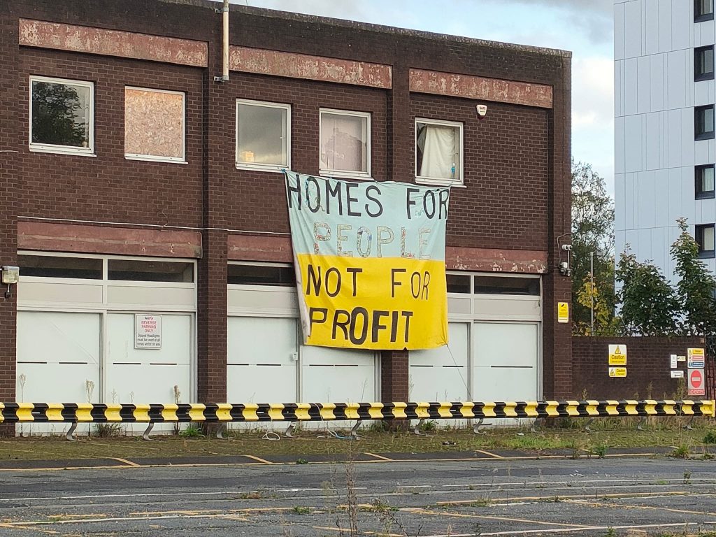 image of a single story red brick building, with a banner hanging that says 'Homes for People, Not for Profit'.