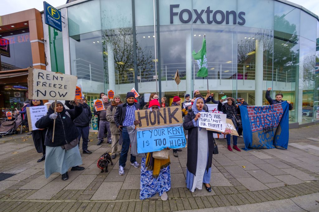 Image shows dozens of protesters outside Foxtons estate agent, one in the foreground holding a sign reading 'honk if your rent is too high!'