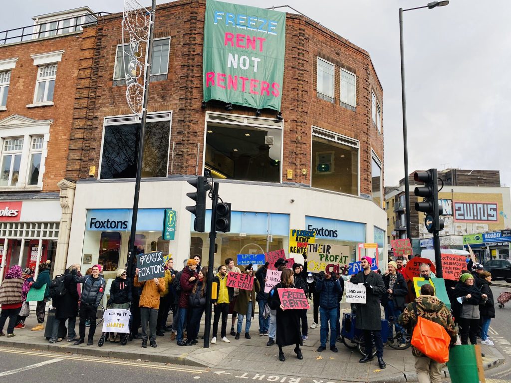 Image shows dozens of protesters outside an estate agent, with a banner dropped over the top of the building reading 'Freeze Rent Not Renters'.