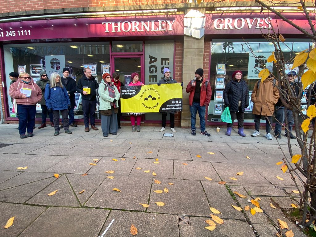 Image shows around 15 housing activists standing outside Thornley Groves estate agent with the Greater Manchester Tenants Union banner. They are wrapped up warm and there are autumn leaves on the ground in the foreground of the picture, the same colour as the yellow banner.