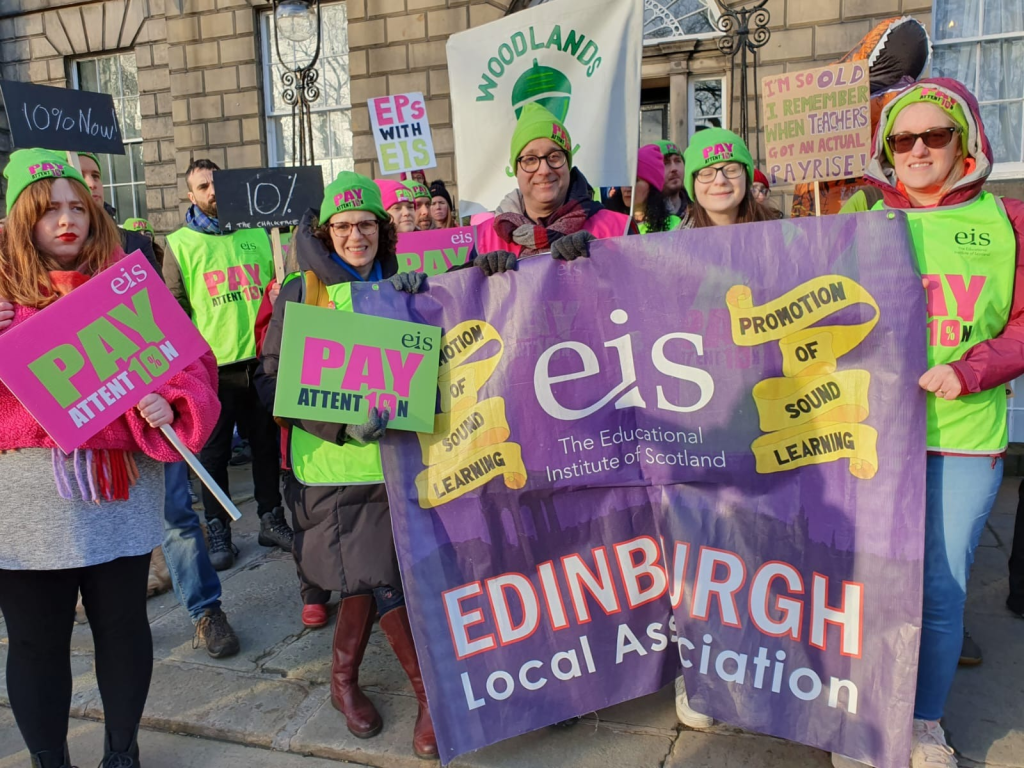teachers in EIS green hats hold banners and placards, saying 'Pay Attent10n', '10% now'.