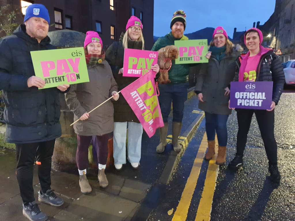 Teachers picket outside a school in Edinburgh, holding placards saying 'Pay Attent10n'. The morning sky is still dark.