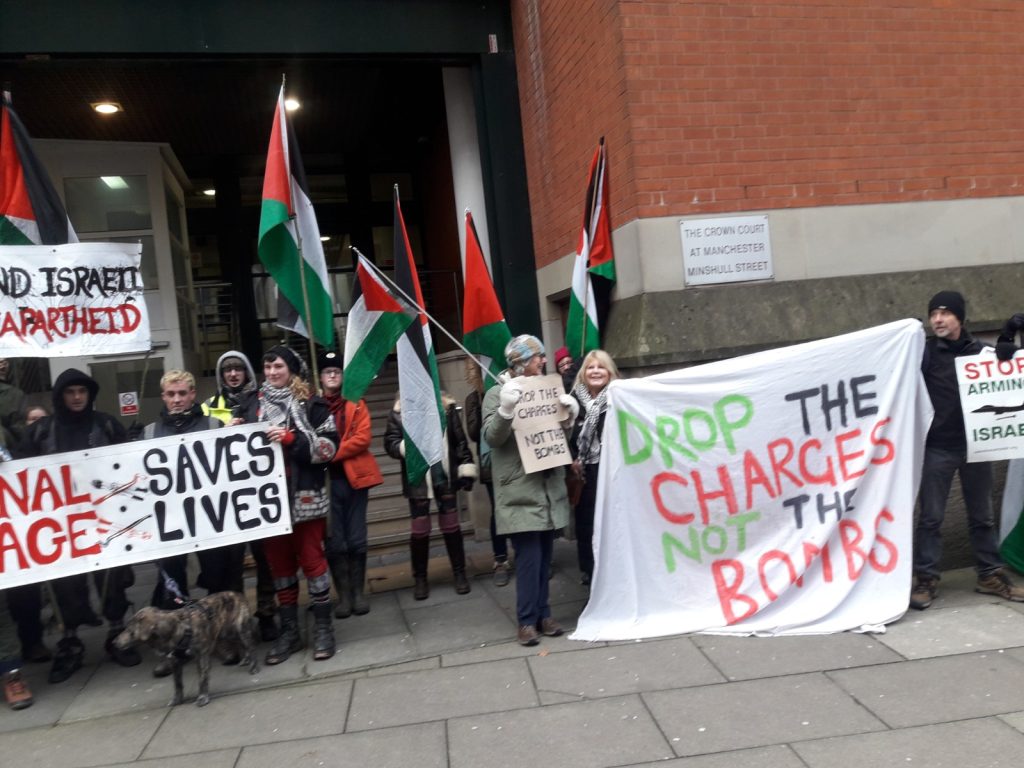 a crowd of mixed ages waves Palestinian flags outside court. Banners in red, green and black writing say 'Drop the charges Not the bombs', 'End Israeli apartheid'.